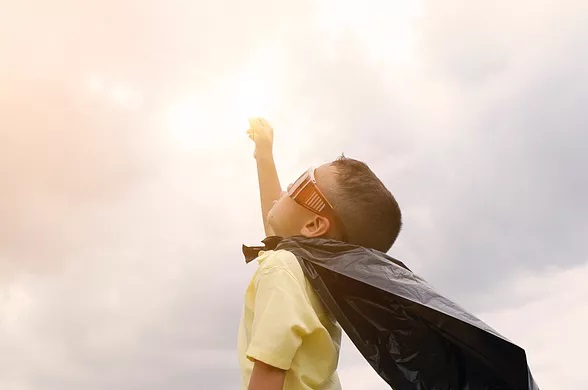A boy wearing sunglasses and a cape is looking up at the sky.
